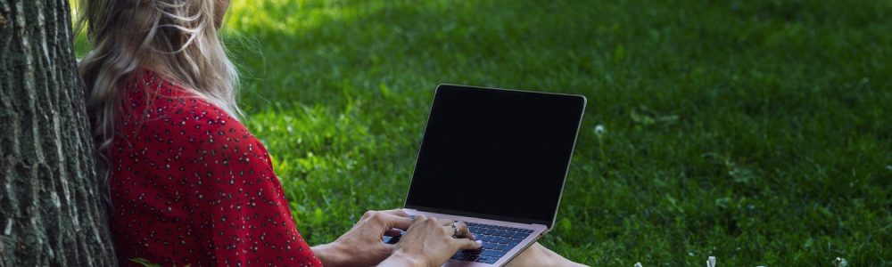 Young woman with laptop on the grass in the park sits by a tree. Remote work and training. Isolated on a black screen. Space for text.